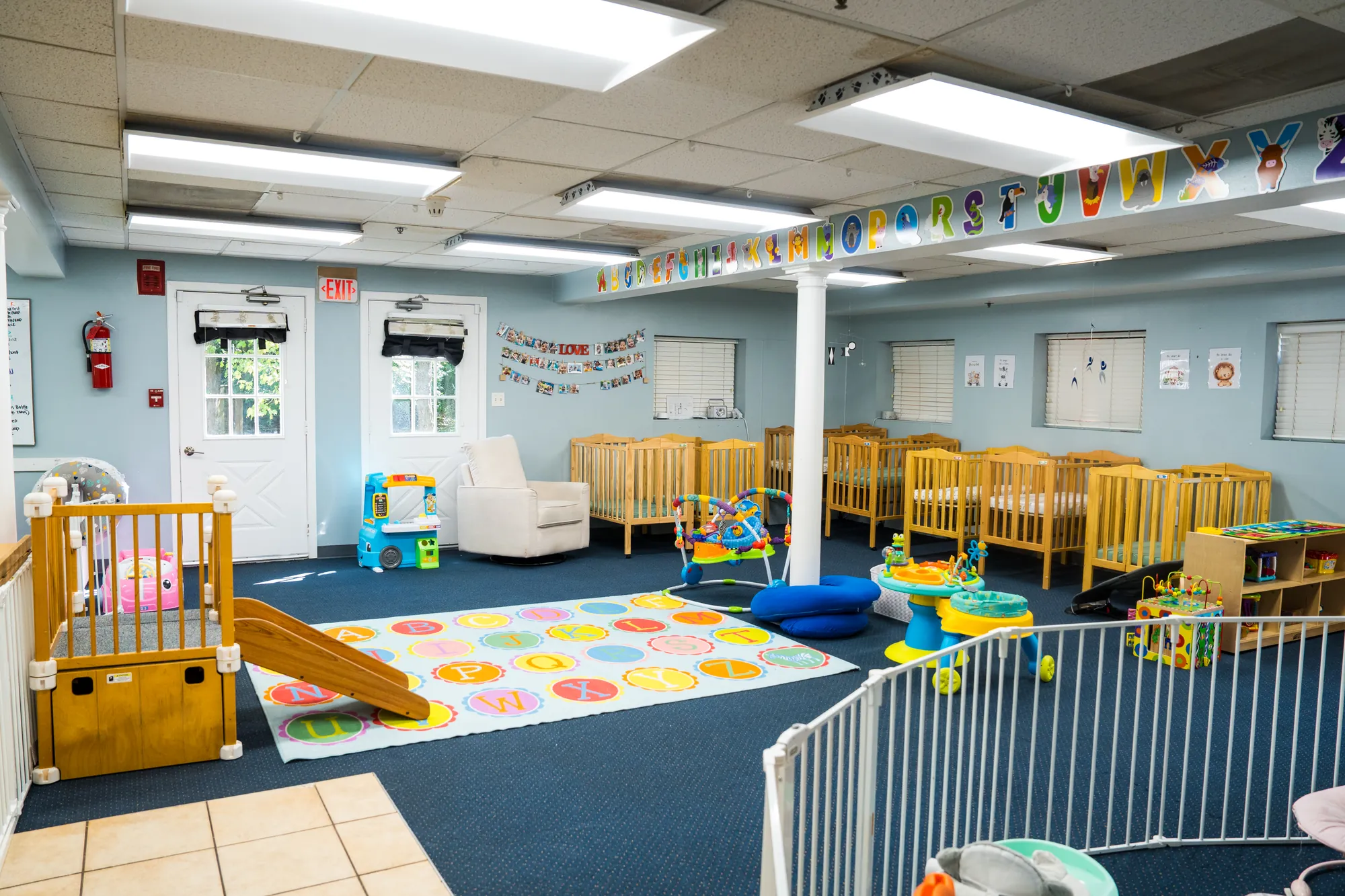 Infant room with rows of cribs, a rocking chair, and an alphabet floor mat