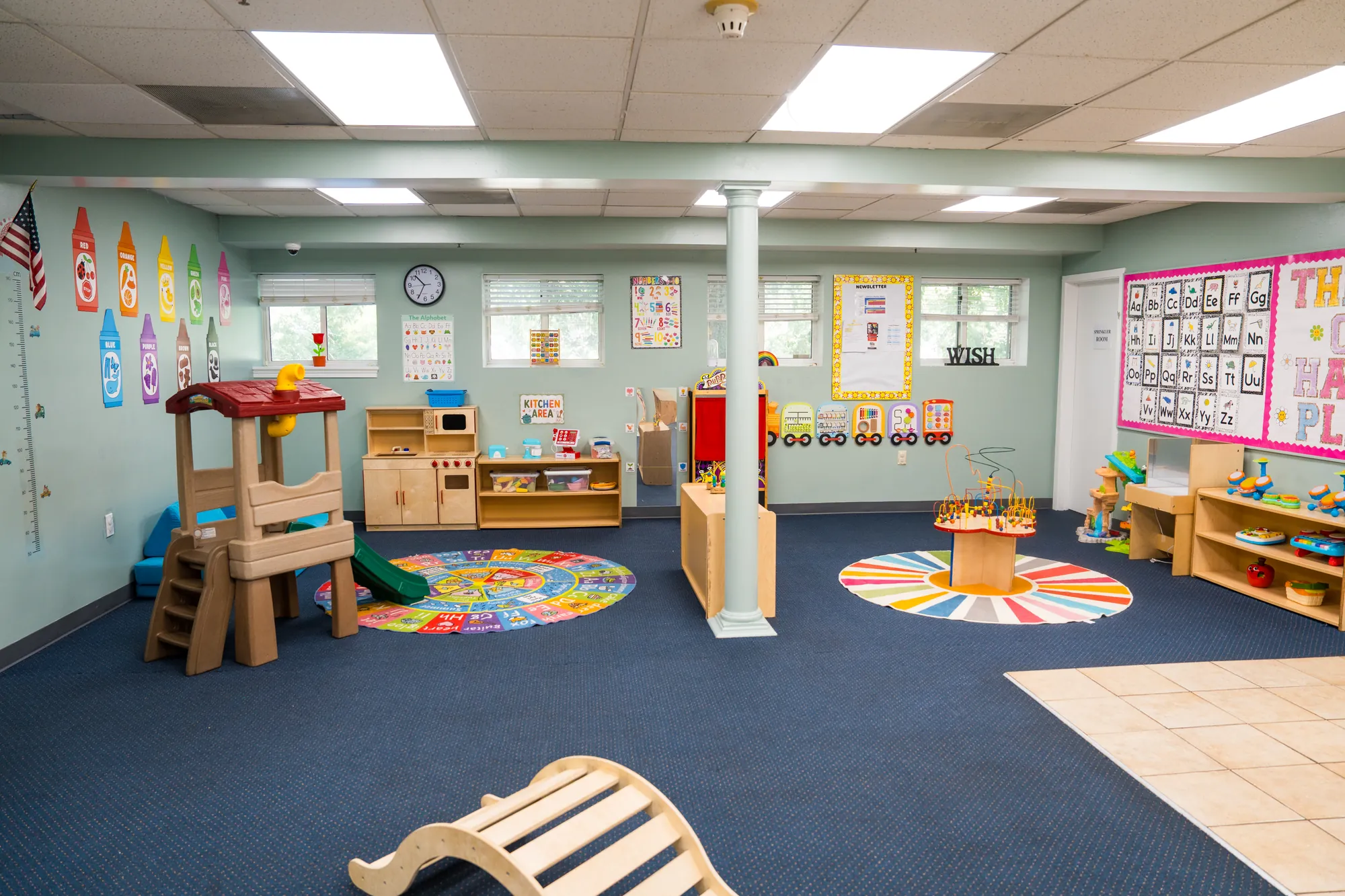 Toddler classroom with a wooden playhouse, activity rugs, and low learning centers